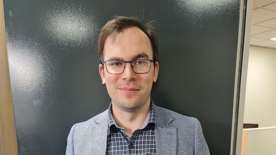 Brendan J Doidge stands in front of a meeting room wearing a suit and glasses