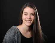 Young woman with long brown hair, black top and grey cardigan smiling at the camera in front of a black background.