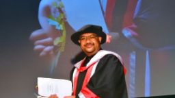 Man is graduation regalia with PhD certificate in hand smiling for the camera