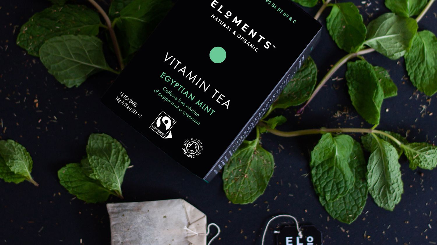 A black box of Eloments Vitamin Tea 'Egyptian Mint' flavour sits on a black stone background atop fresh mint. One tea bag is displayed on the right hand side. 
