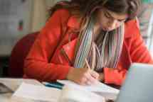 Female student wearing a red coat is writing in her book on a desk