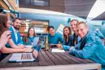 Seven young people sitting around a cafeteria table, smiling to camera.