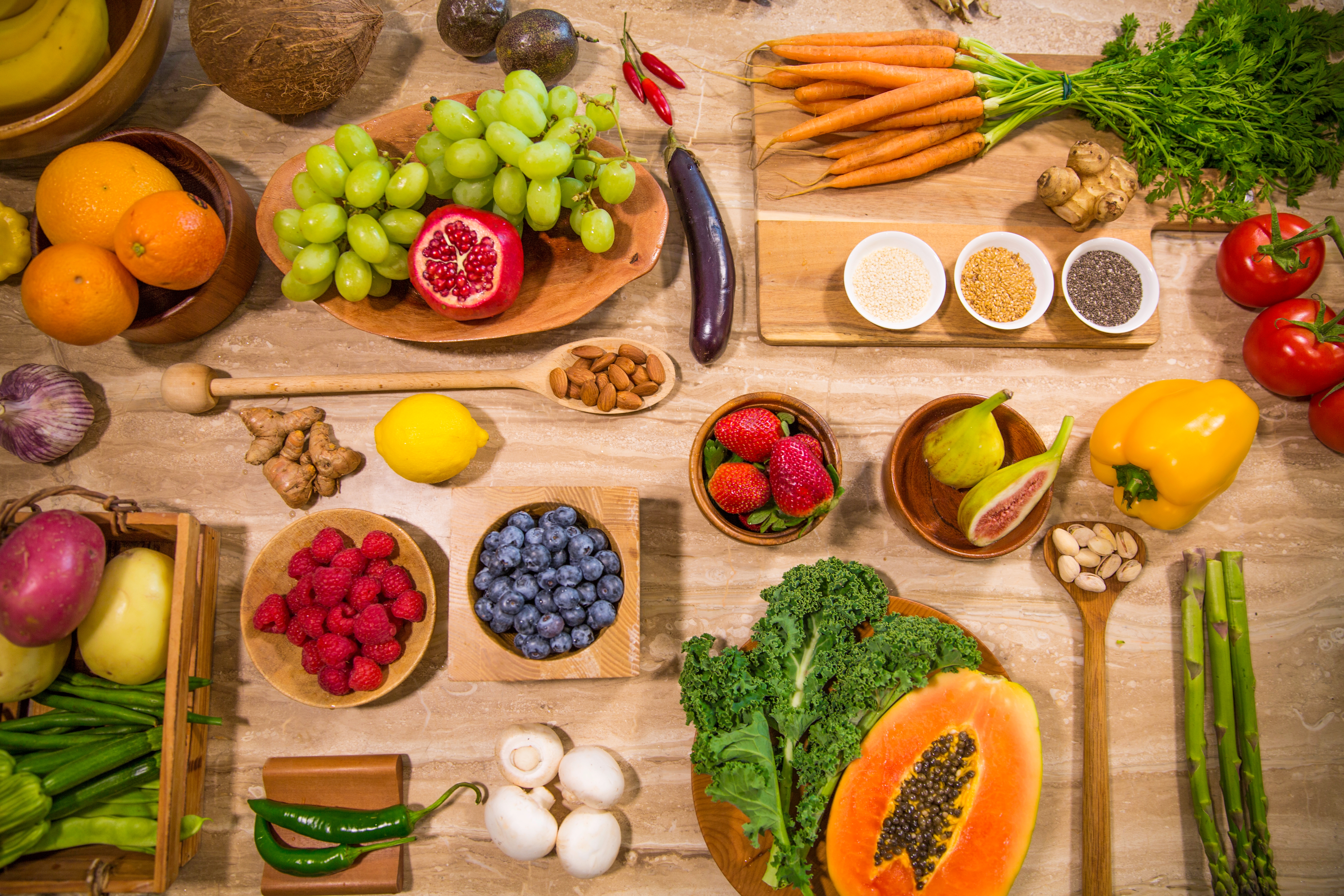 A rainbow of fresh fruit, vegetables, nuts and legumes is enticingly laid out on a wooden surface. 