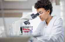 Young female Scientist with short curly black hair working with a microscope in a lab wearing a white lab coat and plastic glasses