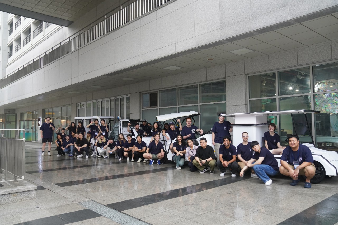 A large group of students and teachers in dark blue shirts crouch by four-wheeled mobility devises, similar to golf buggies