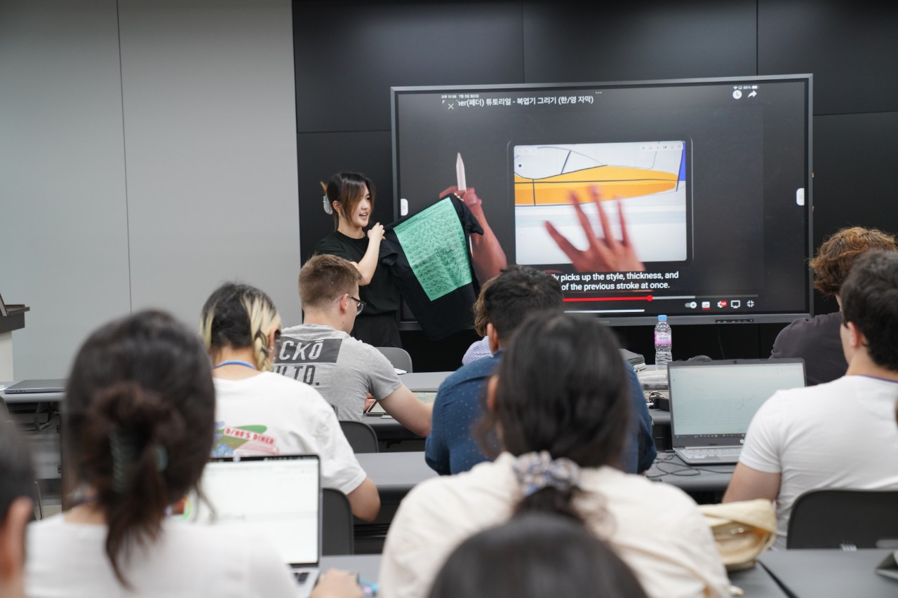 Student sit facing the front of a classroom where someone holds up a t -shirt beside a screan playing a video