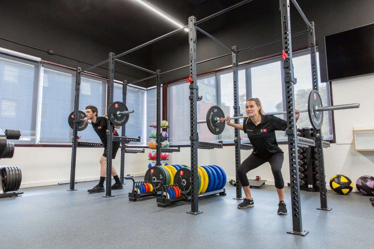 A male and female student wearing black Swinburne apparel using a squat rack