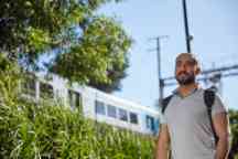 Male student wearing a backpack standing outside with a train passing by in the background.