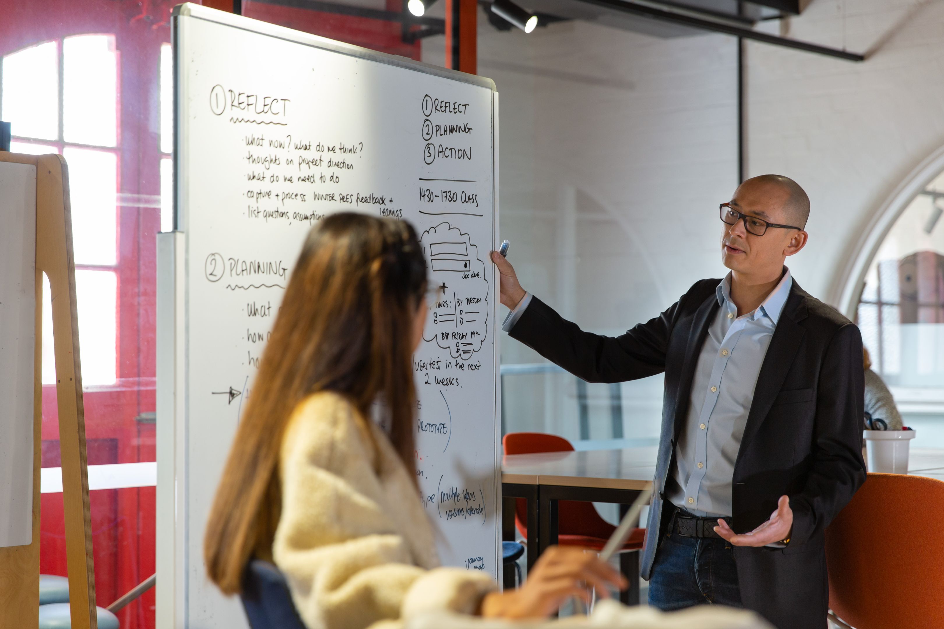 Two Swinburne students are participating in a workshop session. One male student stands next to a whiteboard, discussing a framework with a seated female student in a bright, collaborative classroom.